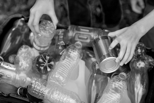 A close-up black and white image showing a person's hands sorting recyclable waste among a collection of plastic bottles and containers. The person's left hand is holding a metal can, while the right hand is reaching to pick up a plastic bottle. The plastic bottles vary in size and shape, some with caps and labels still attached, and are scattered on top of each other. The background is blurred but suggests an outdoor environment, possibly a garden or street setting, with the waste material resting on a flat surface. The scene emphasizes the process of waste segregation, typical of rubbish management tasks carried out by Commercial Waste Removal Kenton, highlighting environmentally conscious disposal methods associated with local rubbish removal services.