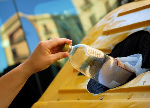A close-up view of a person's hand placing a clear plastic bottle into a yellow waste collection skip on a driveway or street. The bottle appears to be empty, with some labels still visible, and is being inserted into an opening in the skip, which has a black interior. The scene is set outdoors, with a blurred background of a building and blue sky, indicating a typical recycling or rubbish disposal activity likely part of commercial waste removal services in the Kenton area. The skip has a smooth, painted surface, and the person's hand is seen wearing no gloves, suggesting routine handling of waste materials. This image emphasizes the process of waste separation and disposal, aligning with services provided by Commercial Waste Removal Kenton, focused on rubbish collection and management for local businesses or residents.