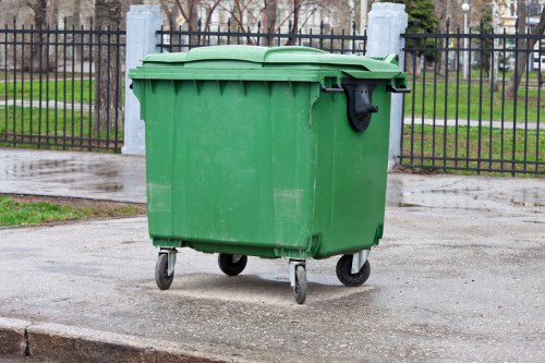 A green plastic wheeled rubbish bin with a closed lid, positioned on a paved area near a black metal fence and a grassy park with trees in the background. The bin appears to be used for waste collection, with a handle on the side and a textured surface. It is situated on a concrete or asphalt surface, with the surrounding environment suggesting an outdoor urban or park setting. The scene is evenly lit, indicating daylight, and the overall appearance is clean and well-maintained, suitable for waste collection services like those provided by Commercial Waste Removal Kenton in the local area of Kenton or nearby postcode regions.
