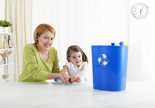 The image shows a woman and a young girl sitting at a white kitchen or dining table, smiling and engaging with a clear plastic bag of waste or recyclables. The woman, with short blonde hair and wearing a lime green long-sleeved top, is positioned on the left side of the image, while the girl, with dark hair tied in pigtails and dressed in a white top, is seated beside her on the right. In front of them, to the right side of the table, there is a large, upright blue recycling bin with a white recycling symbol on its front. The background features a bright, well-lit room with white walls, a wall clock showing approximately 12:08, and a white shelf unit with small decorative items. The scene suggests an emphasis on household waste management and recycling, which relates to the services provided by Commercial Waste Removal Kenton in managing general rubbish and waste collection locally in the Kenton area.