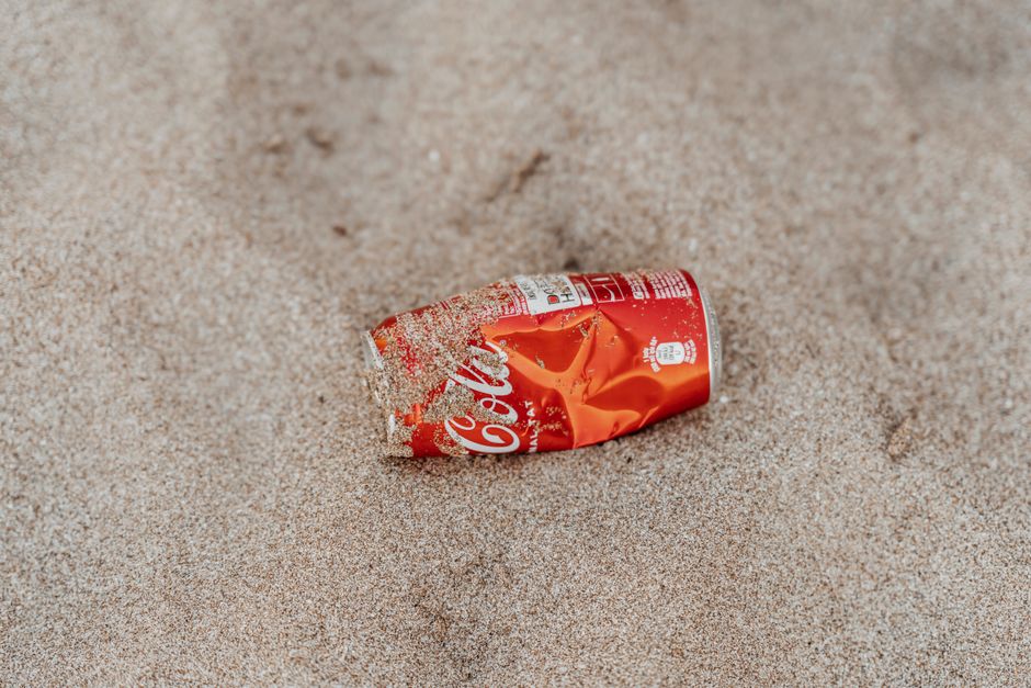 A crumpled and partially buried red aluminium can of Coca-Cola lies on a sandy surface, with the word 'Coca-Cola' visible on its side. The can appears damaged, with dents and tears, and is coated with