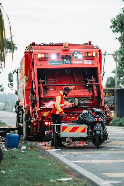 A person with curly dark hair wearing a black t-shirt and checkered shorts is standing on a paved sidewalk next to two large, open-top skips filled with construction or renovation waste, including bro